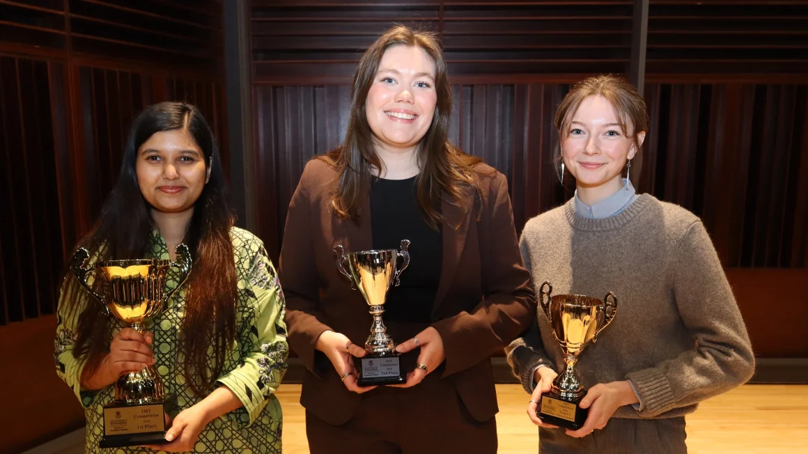 Three female students smile on a stage. Each holds a small gold trophy.