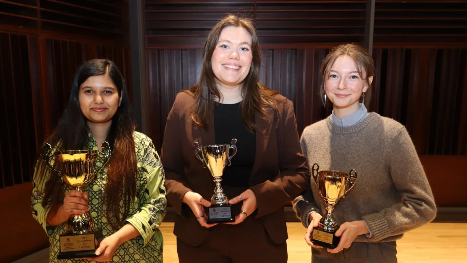 Three female students smile on a stage. Each holds a small gold trophy.