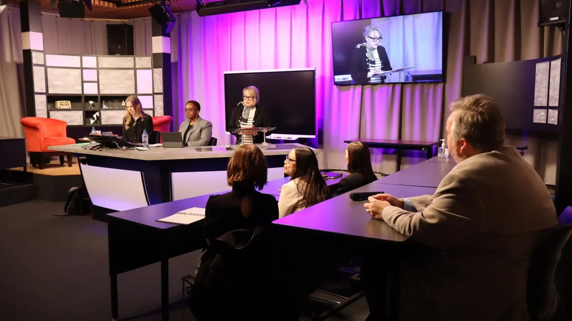 Panelists speak on stage in front of a purple background with audience members in the foreground.