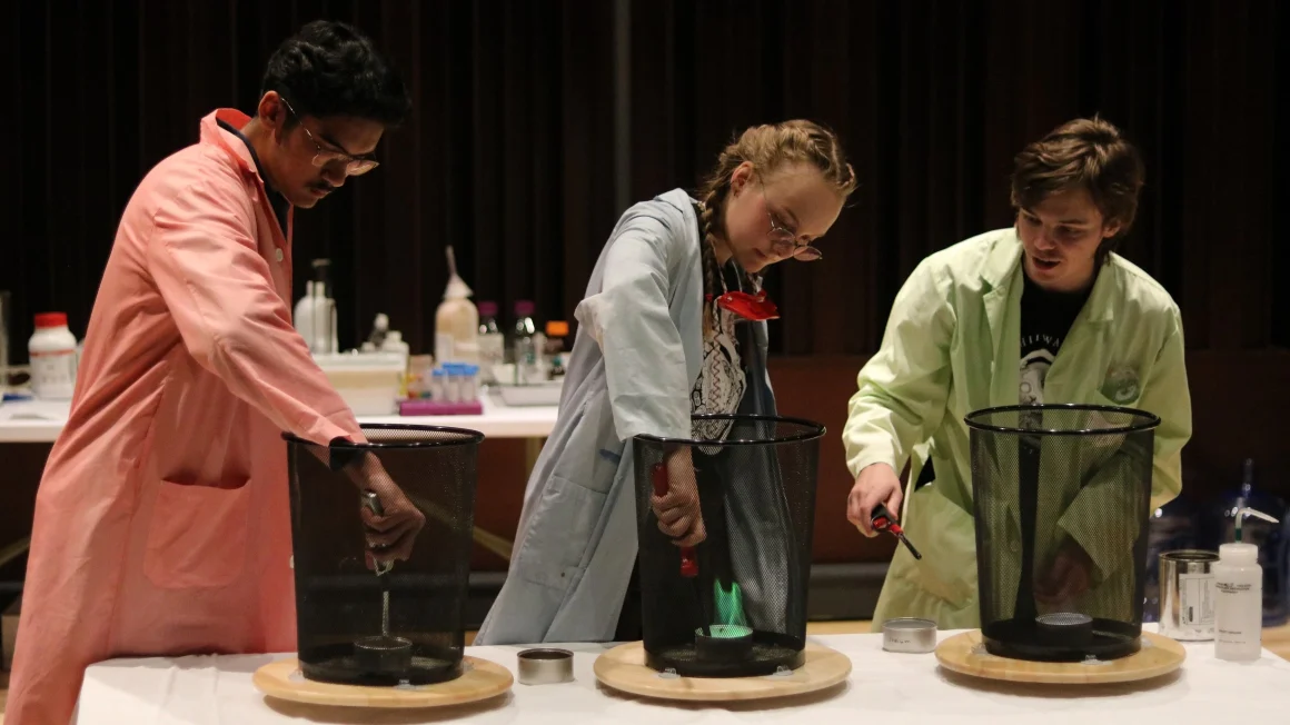 Three student assistants in lab coats produce small flames in a controlled tabletop chemistry experiment for children.