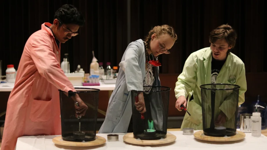 Three student assistants in lab coats produce small flames in a controlled tabletop chemistry experiment for children.