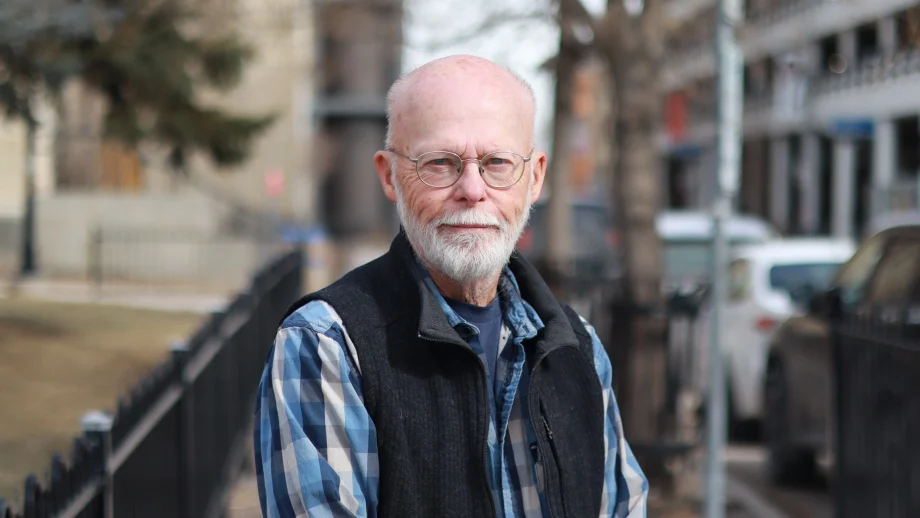 A male professor in a blue plaid shirt and black fleece vest stands on a campus sidewalk on a cloudy day.