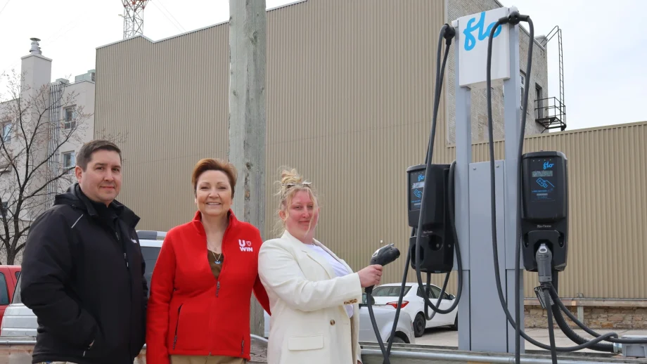 A man in a black jacket, a woman in a red fleece, and a woman in a white blazer stand next to an electrical vehicle charging station in a surface parking lot on campus.