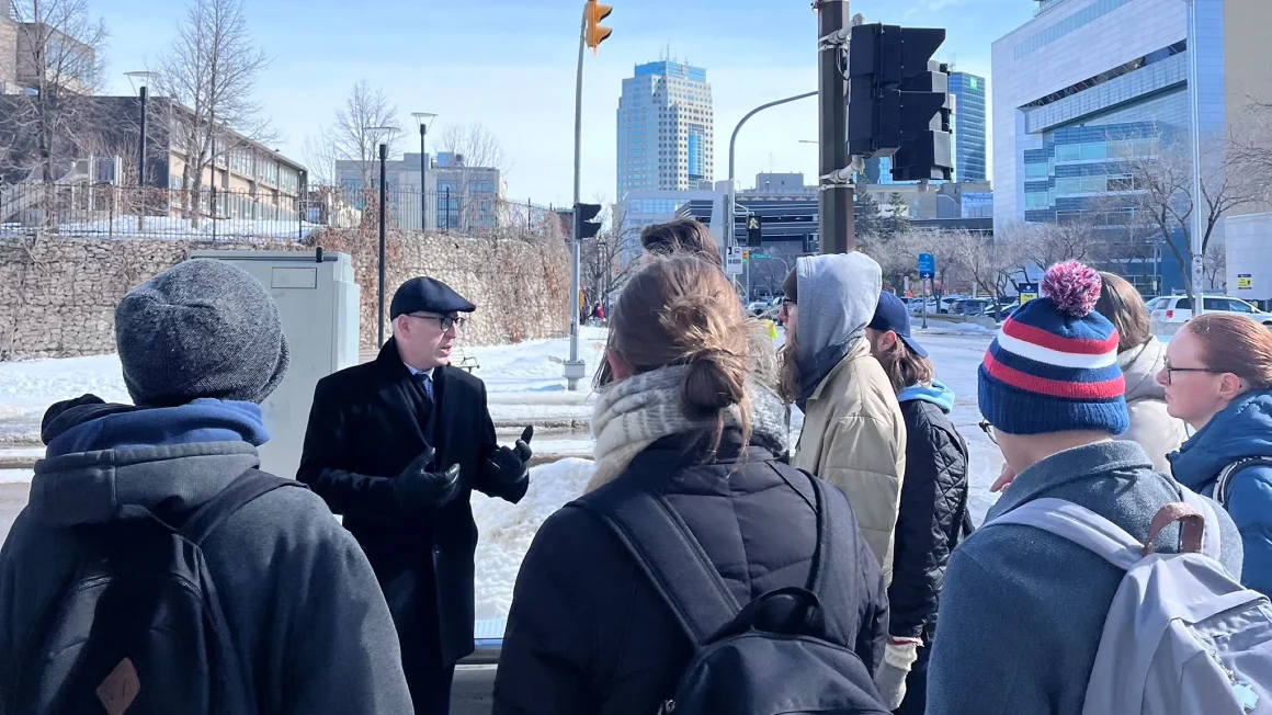 Dr. Jino Distasio, in a black coat and hat, speaks to a group of students outside.