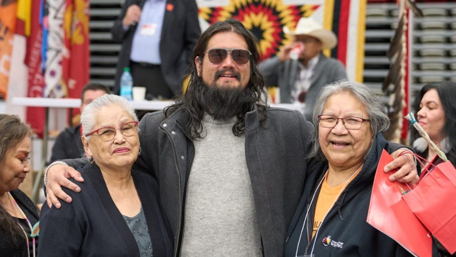 A male graduate wearing sunglasses and a black jacket is embraced by two elders.