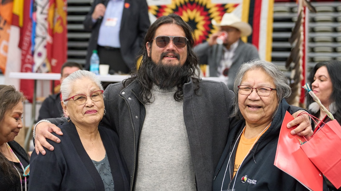 A male graduate wearing sunglasses and a black jacket is embraced by two elders.