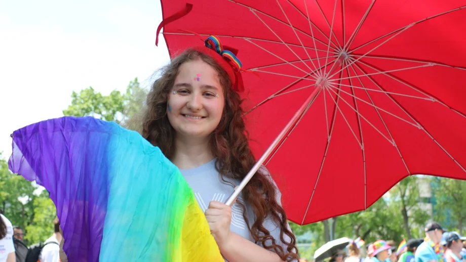 A young person with long brown hair holds a red umbrella and a rainbow-coloured fan at an outdoor Pride celebration.