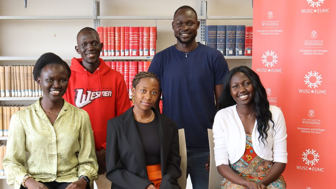 Monica Ayuen Dot, Sunday Emmanuel, Harriet Kabaosomi, Peter Ajang Deng, and Nyamal Kuon are standing in front of a stack of books in the library.