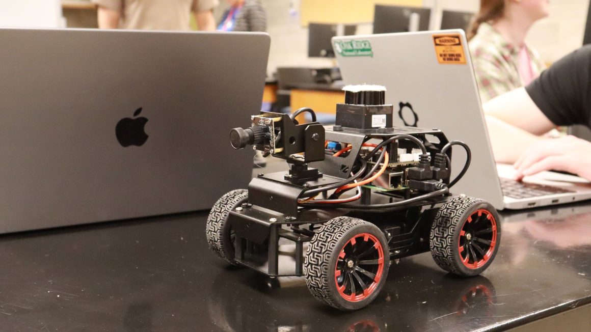 A motorized four-wheeled robot the size of a small microwave sits on a table next to laptops in a classroom.