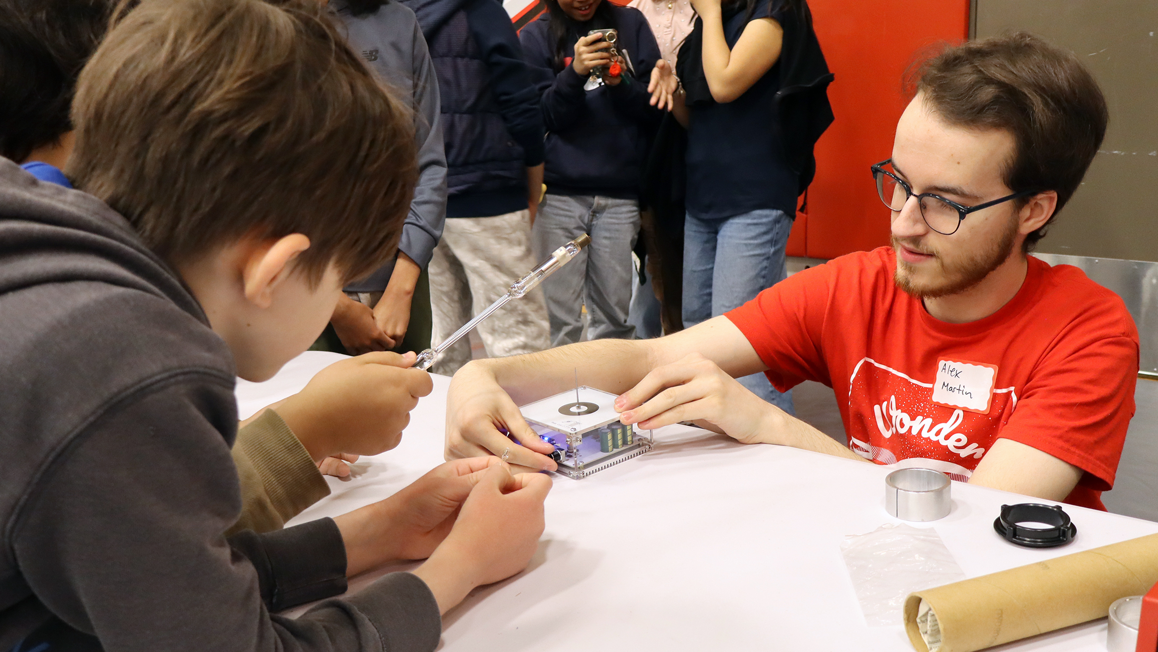 Person adjusts scientific equipment on a table, while children around watch.