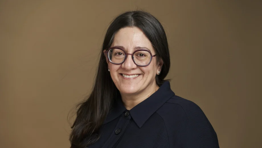 A portrait of a female professor with brown hair and glasses smiling in front of a tan photo backdrop.