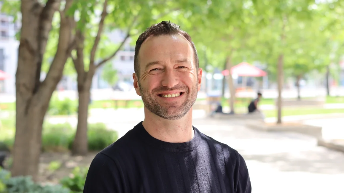A man with brown hair and a black shirt smiles as he stands under some trees in the shade.