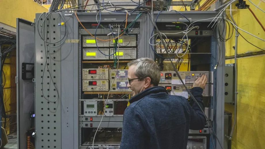 A physics professor stands in front of a shelving unit holding lab equipment and wires.