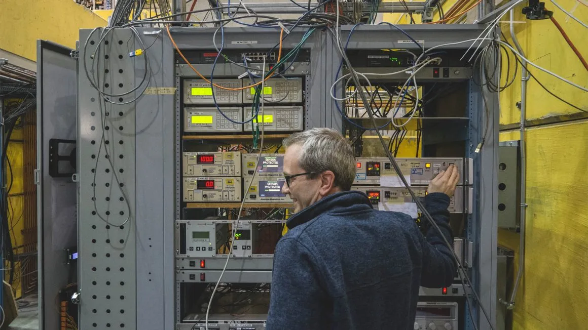 A physics professor stands in front of a shelving unit holding lab equipment and wires.