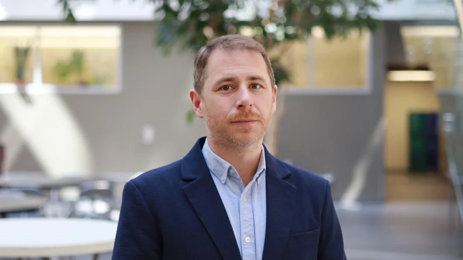A man in a light blue shirt and dark blue blazer stands in a sunny atrium.