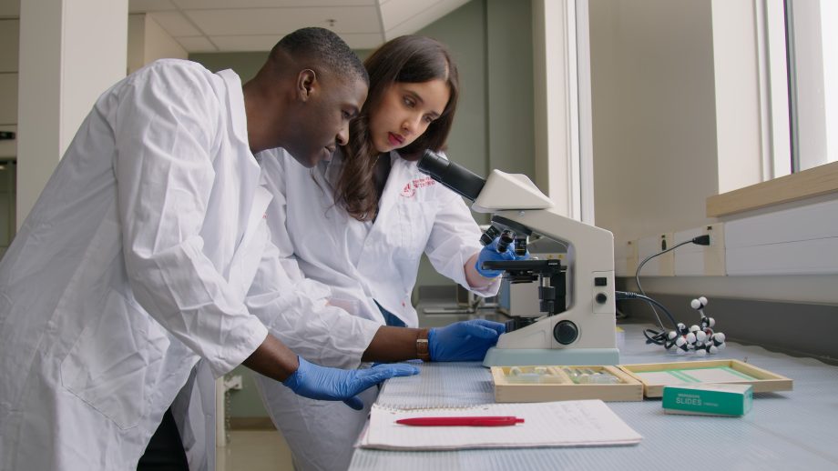Two students in white lab coats use a tabletop microscope in a lab.