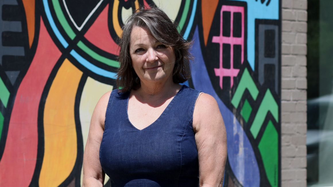 Dr. Shauna MacKinnon is standing in front of an Indigenous mural at UWinnipeg's Selkirk campus