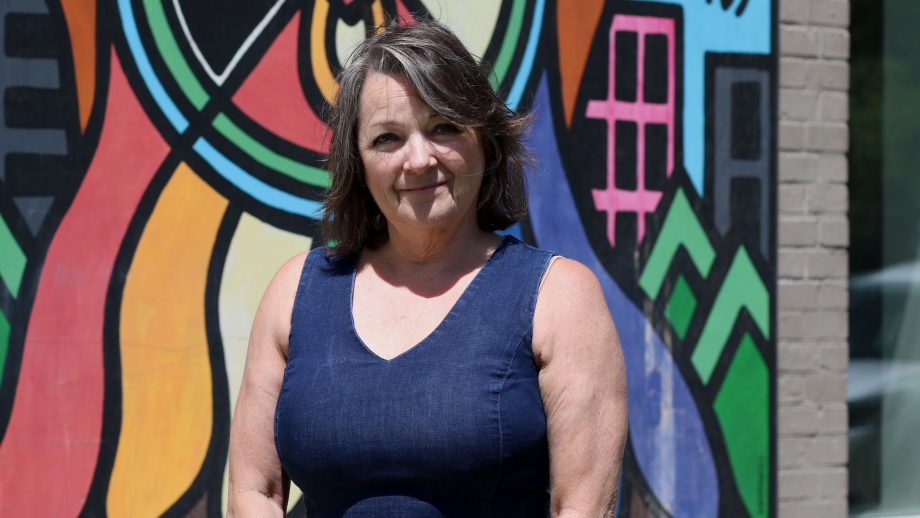 Dr. Shauna MacKinnon is standing in front of an Indigenous mural at UWinnipeg's Selkirk campus