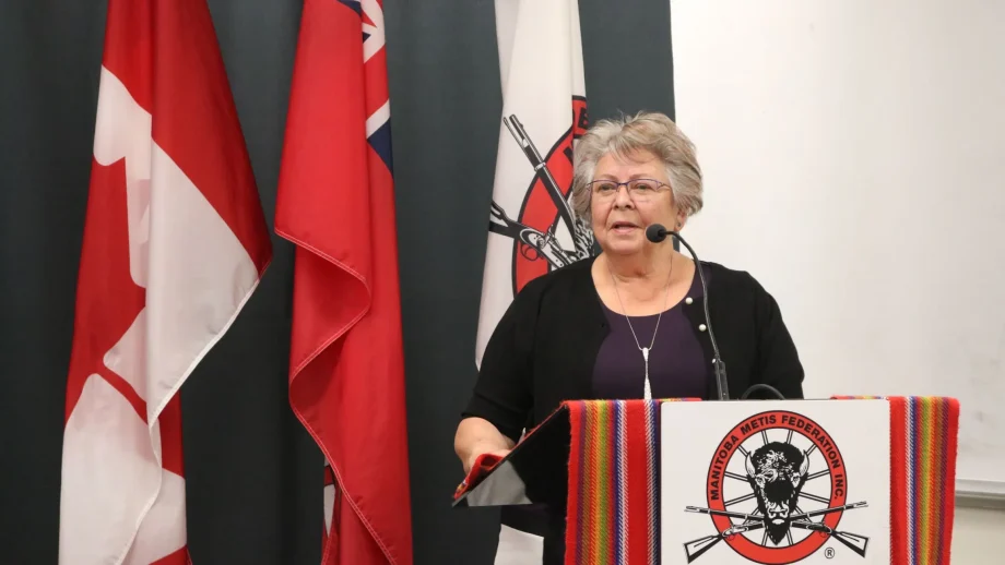 Joan Ledoux in a black shirt speaking at a podium with flags beside her.