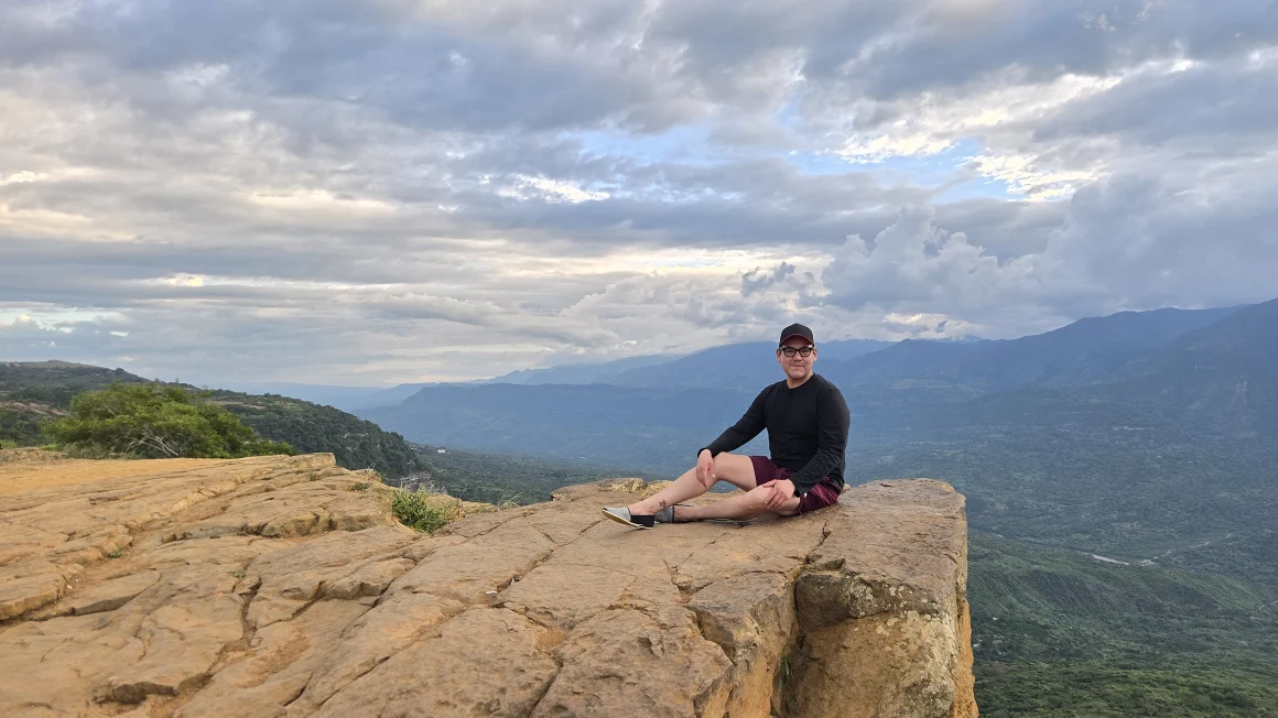 person sitting near the edge of a large rock formation with sky and clouds in the background
