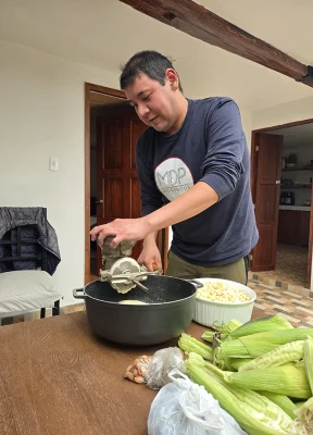 Person using a kitchen tool over a large pot. Cobs of corn are on the table.