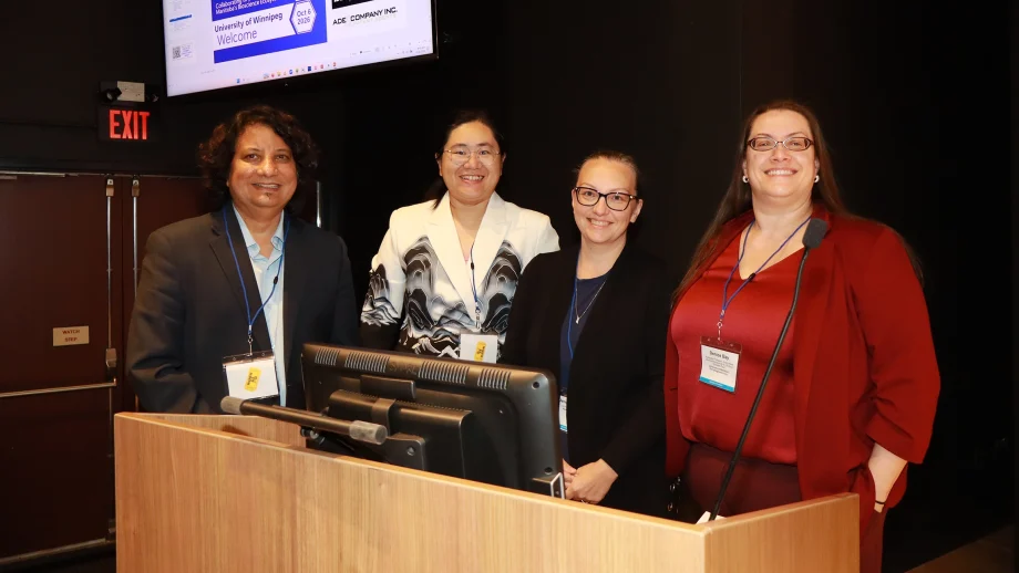 Four people stand behind a podium. A screen is behind them in the upper left side of the photo.